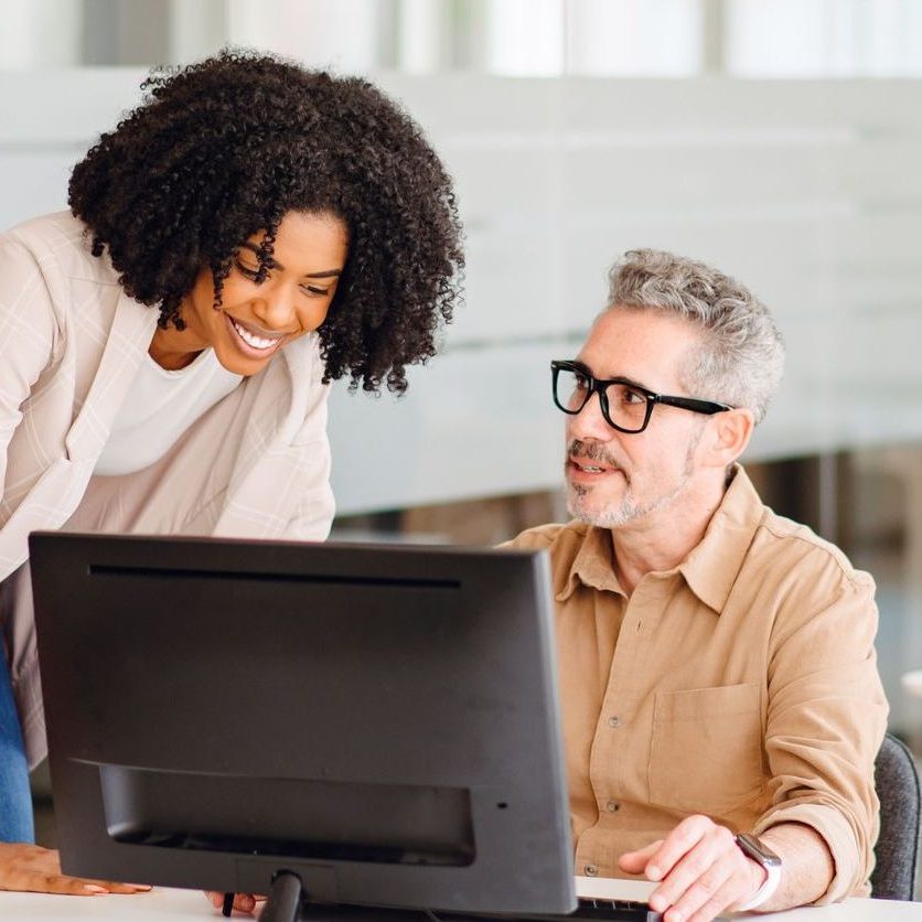 Cheerful woman in business casual attire leaning over a male colleague, sharing ideas or advice Colleagues working together on district-wide email communication