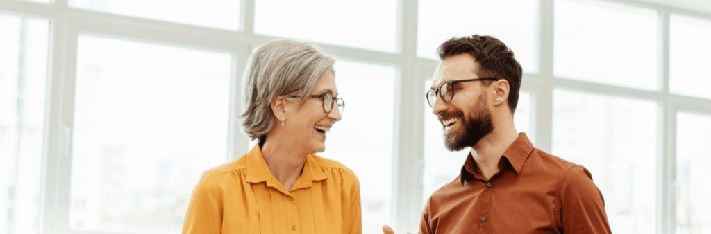 A female in a yellow shirt is smiling and discussing work with a male colleague in a dark orange shirt.