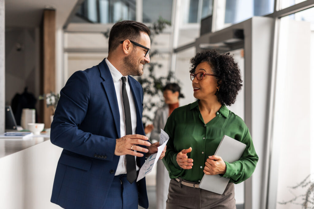 Two business professionals are smiling, talking and walking through a bright modern office building, holding documents and a laptop, engaging in a productive conversation