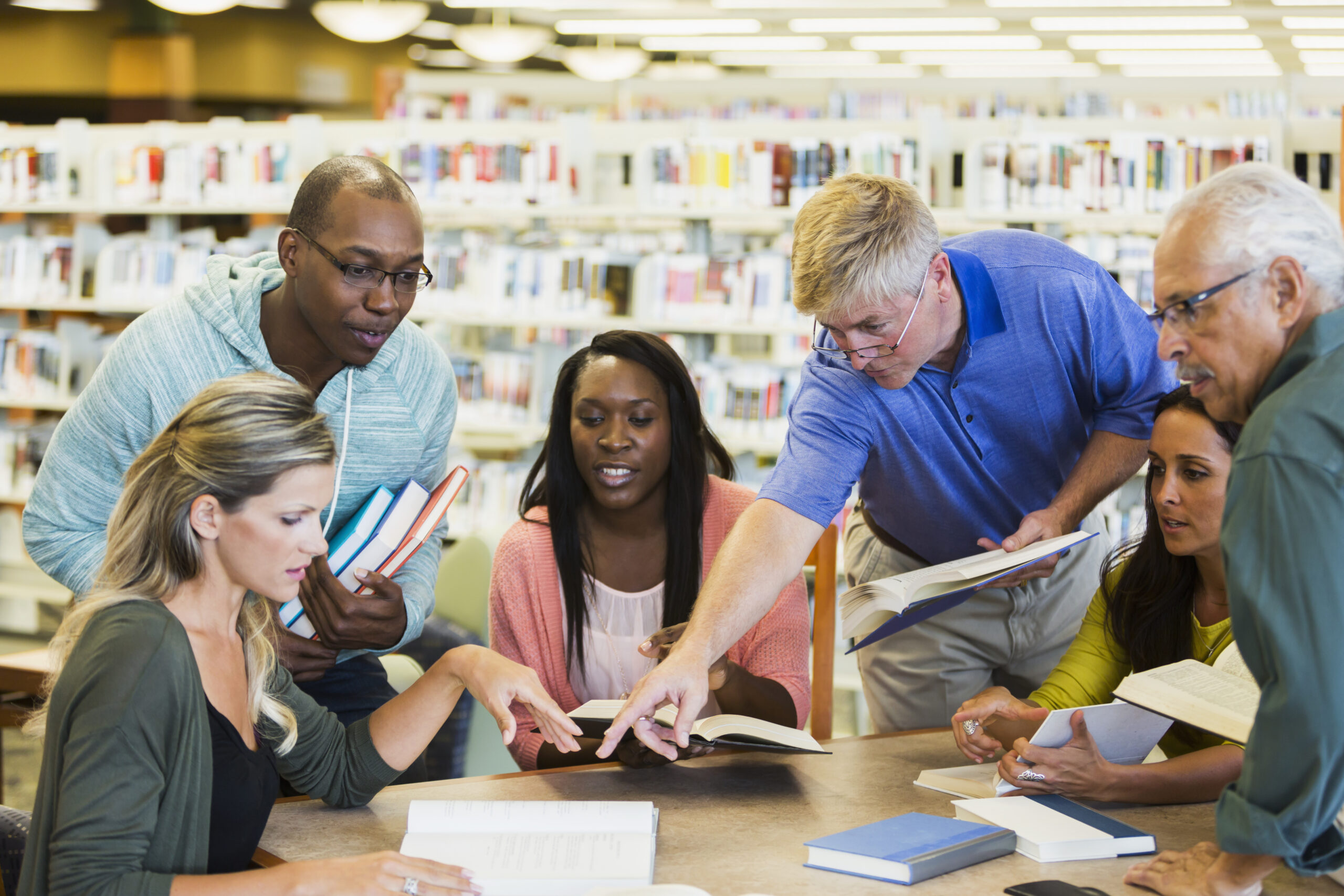 Graduate school or continuing education: a multi-ethnic group of adult students meeting and studying together in the library. Mixed ages, 20s to 70s. They are all sitting or standing around a table holding books. Everyone is looking at the book that a man in a blue shirt is pointing toward.